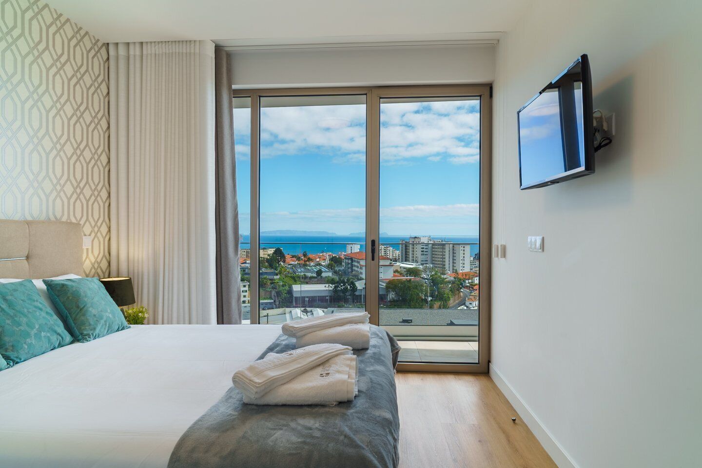 A hotel room with a large bed, a TV mounted on the wall, and a view of the city and sea through the window.