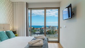 A hotel room with a large bed, a TV mounted on the wall, and a view of the city and sea through the window. - GuestReady - The perfect place to relax in Funchal (Funchal)