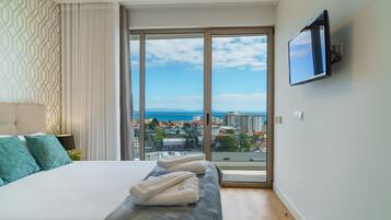 A hotel room with a large bed, a TV mounted on the wall, and a view of the city and sea through the window.