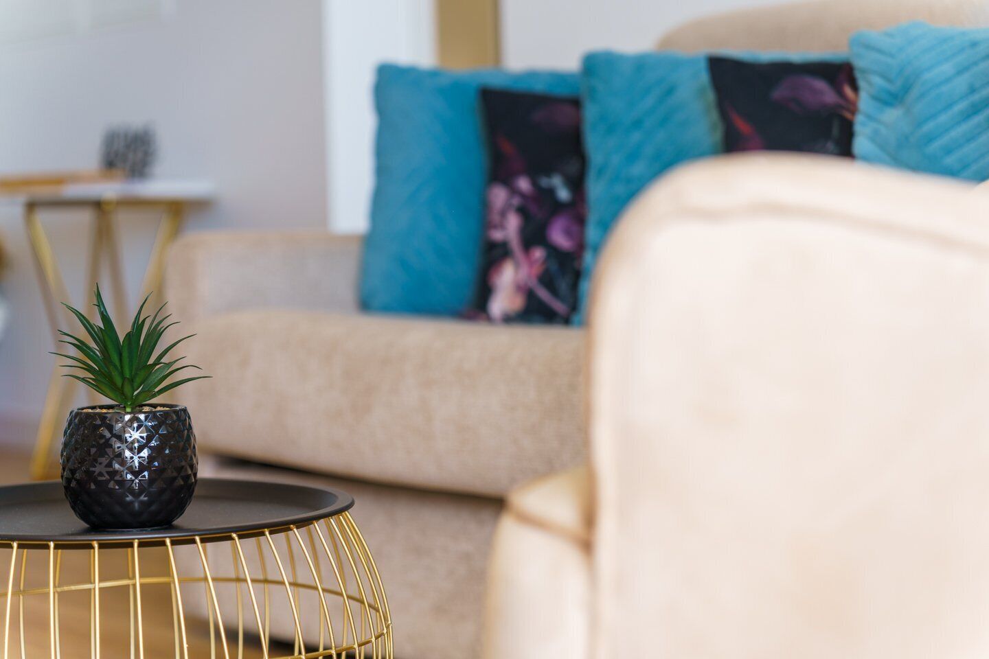A living room with a beige sofa, a small round table with a plant, and a gold wireframe table.