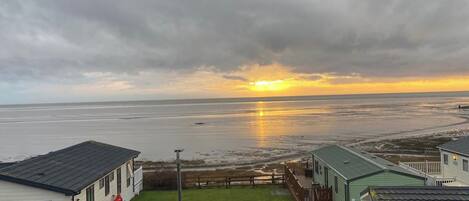 A view of a beachside property with a grassy lawn, a wooden deck, and a clear view of the ocean at sunset.