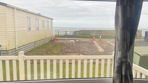 A view from a window overlooking a grassy area with a white picket fence, a mobile home, and a distant beach. - Seaside Retreat-Sleeps 8-Free Parking (Morecambe)