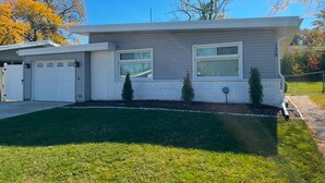 A modern single-story house with a garage, a well-maintained lawn, and mature trees in the background. - Charming 3-bedroom house in Park Forest with WiFi, Large Flat Panel TVs & Google (Park Forest)