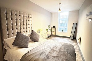 A bedroom with a tufted headboard, a bed with two pillows, a bedside table with a lamp, a window with blinds, and a mirror on the wall.