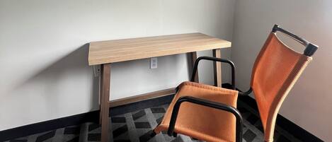 A corner room with a desk, an orange office chair, a patterned carpet, and a framed abstract painting on the wall.