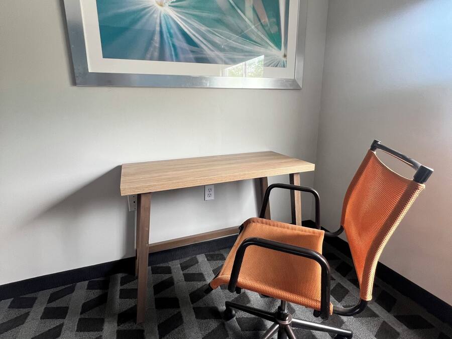 A corner room with a desk, an orange office chair, a patterned carpet, and a framed abstract painting on the wall.