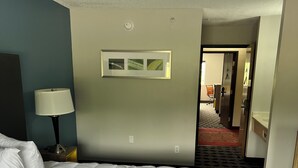 A hotel room with a large bed, a nightstand with a lamp, a framed picture on the wall, and a door leading to a hallway. - LakeWood Inn & Suites (Dowagiac)