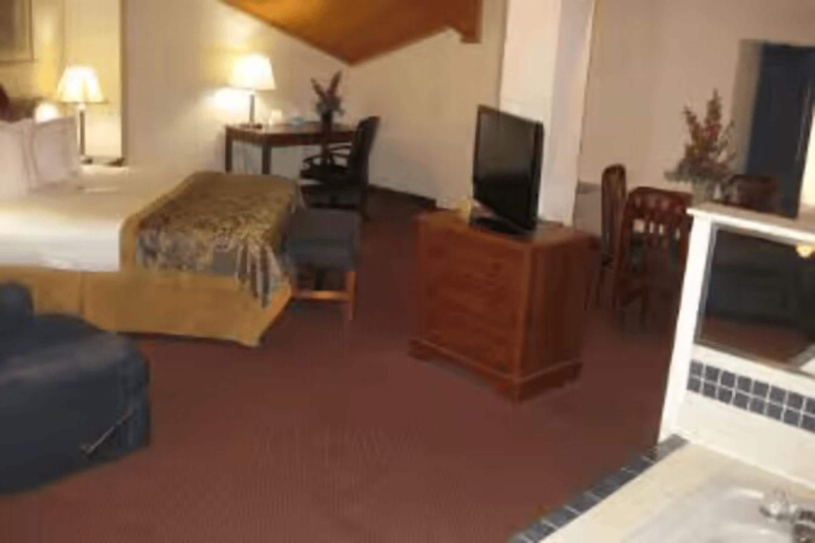 A hotel room with a bed, a television on a wooden stand, a desk with a chair, and a small table with a lamp.