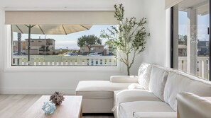 Uma sala de estar moderna com um sofá, uma mesinha com objetos decorativos, uma janela grande com vista para edifícios e uma rua, e um vaso com planta.