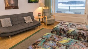 A cozy room with a grey sofa, a small table with a lamp, a chair, and two beds with colorful bedding.