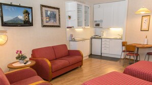 Une pièce compacte comprenant un canapé, deux lits simples, une petite table avec une plante, un coin cuisine et un téléviseur fixé au mur.