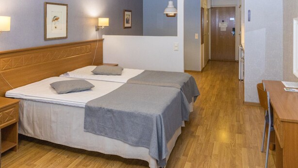 A hotel room with a double bed, bedside tables, a desk, and a chair. There are framed pictures on the wall and a hanging light fixture.