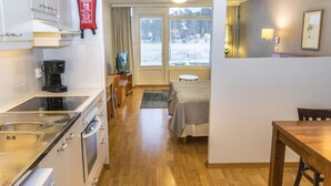 A compact hotel room with a kitchenette, a bed, a small table, and a window with a view of a snowy landscape.