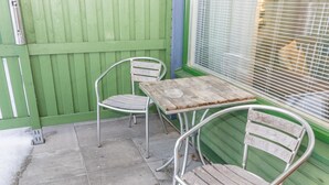 A patio with a metal table and two metal chairs, a green fence, and a window with blinds.