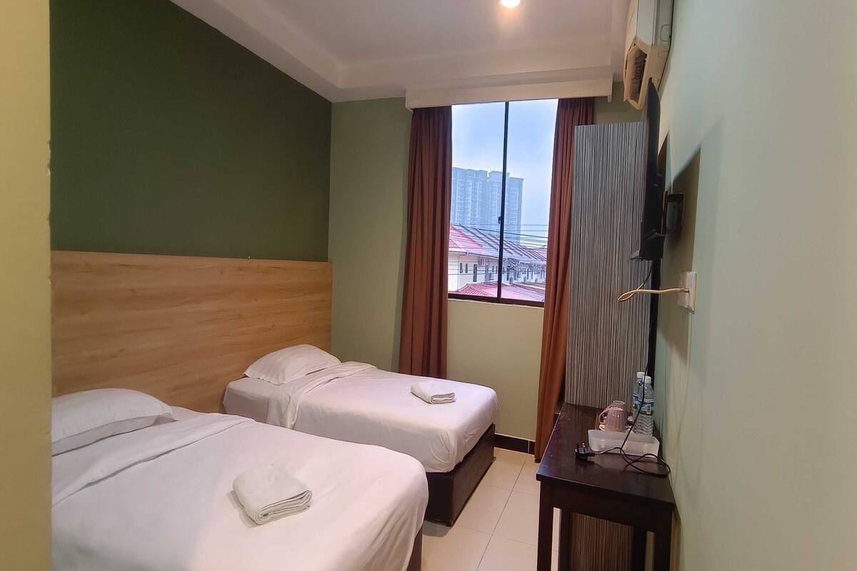 Habitación de hotel con dos camas individuales, cabecera de madera, una mesita con hervidor y vistas a edificios a través de una ventana.