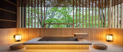 Un espacio de spa de madera con un jacuzzi, dos luces fijadas a la pared y vistas a árboles a través de persianas de bambú.