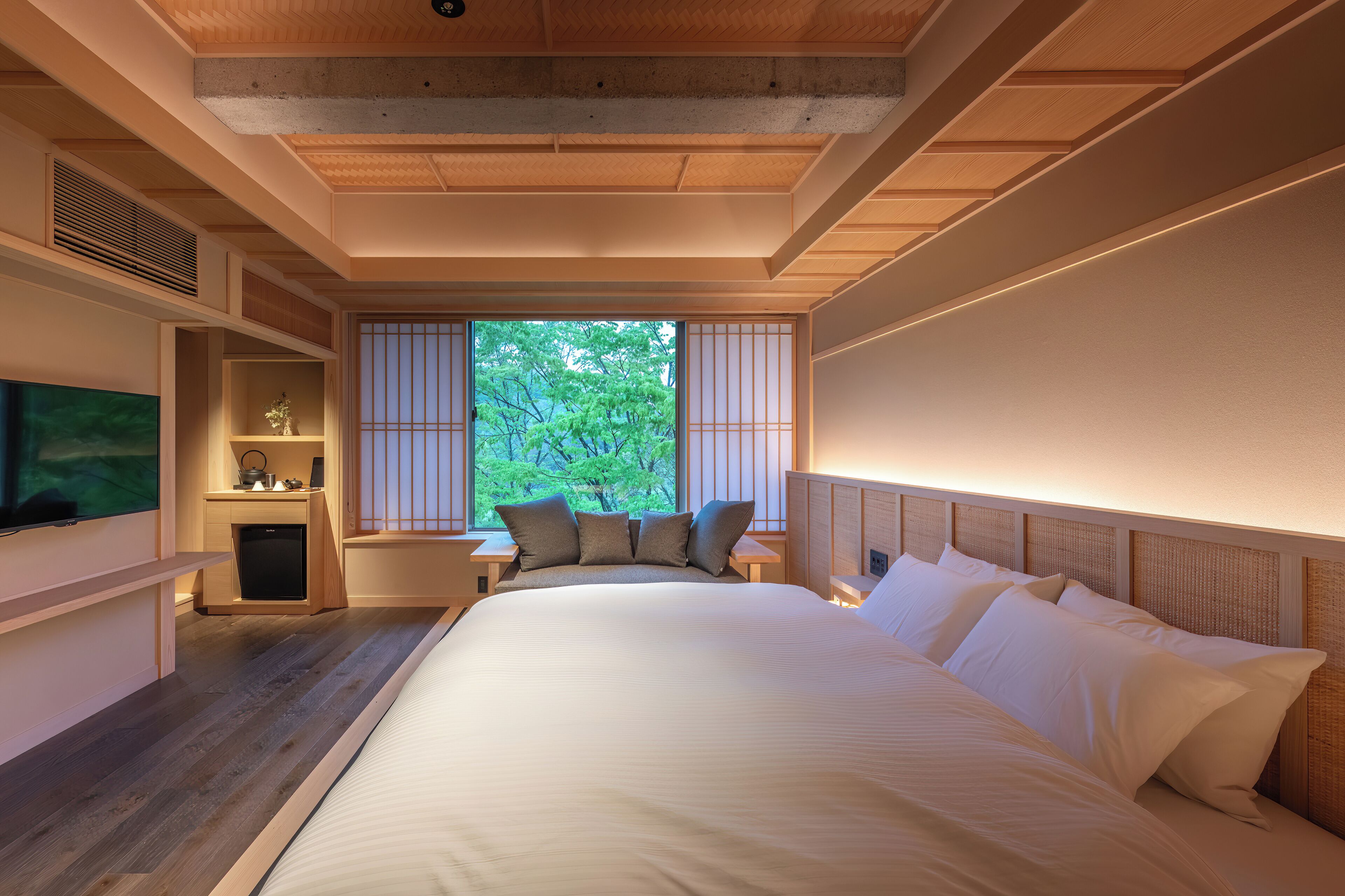Een slaapkamer met een groot bed, houten plafond, ingebouwde planken, een televisie en een raam met uitzicht op bomen.
