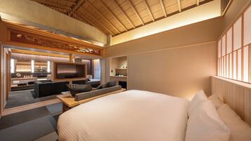Una habitación de hotel moderna con una cama grande, un televisor, una zona de estar con sofá y sillones, y un techo de madera.