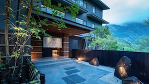 A modern building with a stone pathway leading to a wooden entrance, surrounded by greenery and a mountainous backdrop. - Nazuna Hakone Miyanoshita (Hakone)
