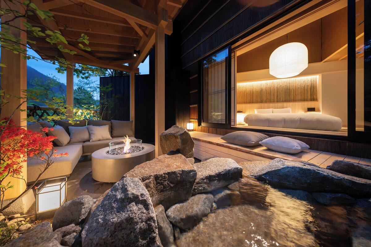 A modern outdoor seating area with a fire feature, a water stream, and a view of a well-lit interior room.