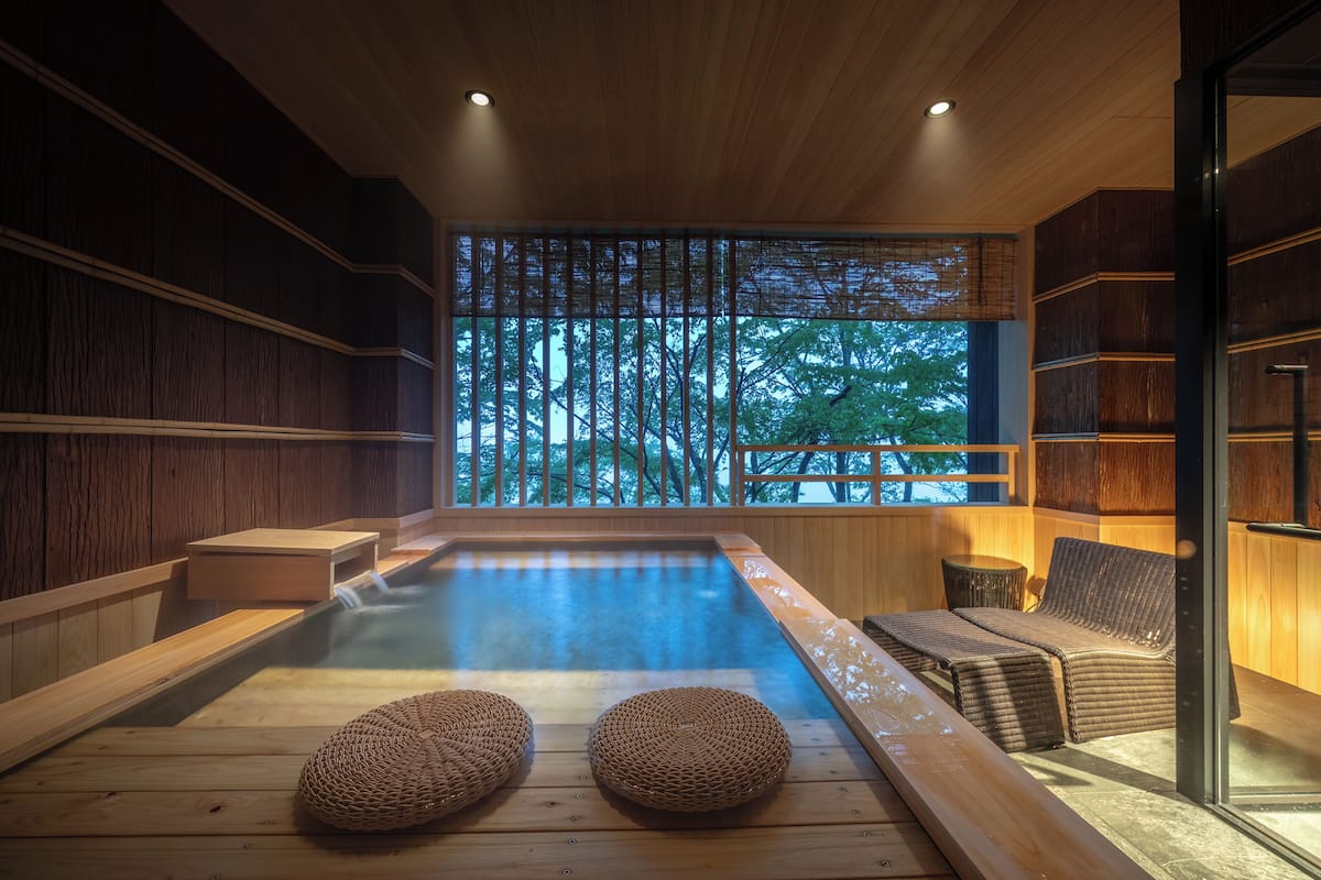 A wooden interior with a hot tub, woven seating, and a large window with a view of trees.