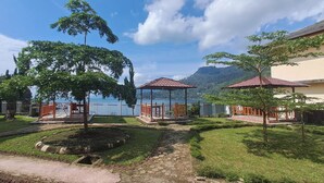 Un gazebo con tetto in tegole, immerso in un’area erbosa con alberi e vista su un lago e le montagne.