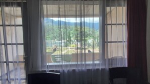 Una stanza con un'ampia finestra, tende leggere, due sedie e vista su un giardino curato con alberi e recinzione.