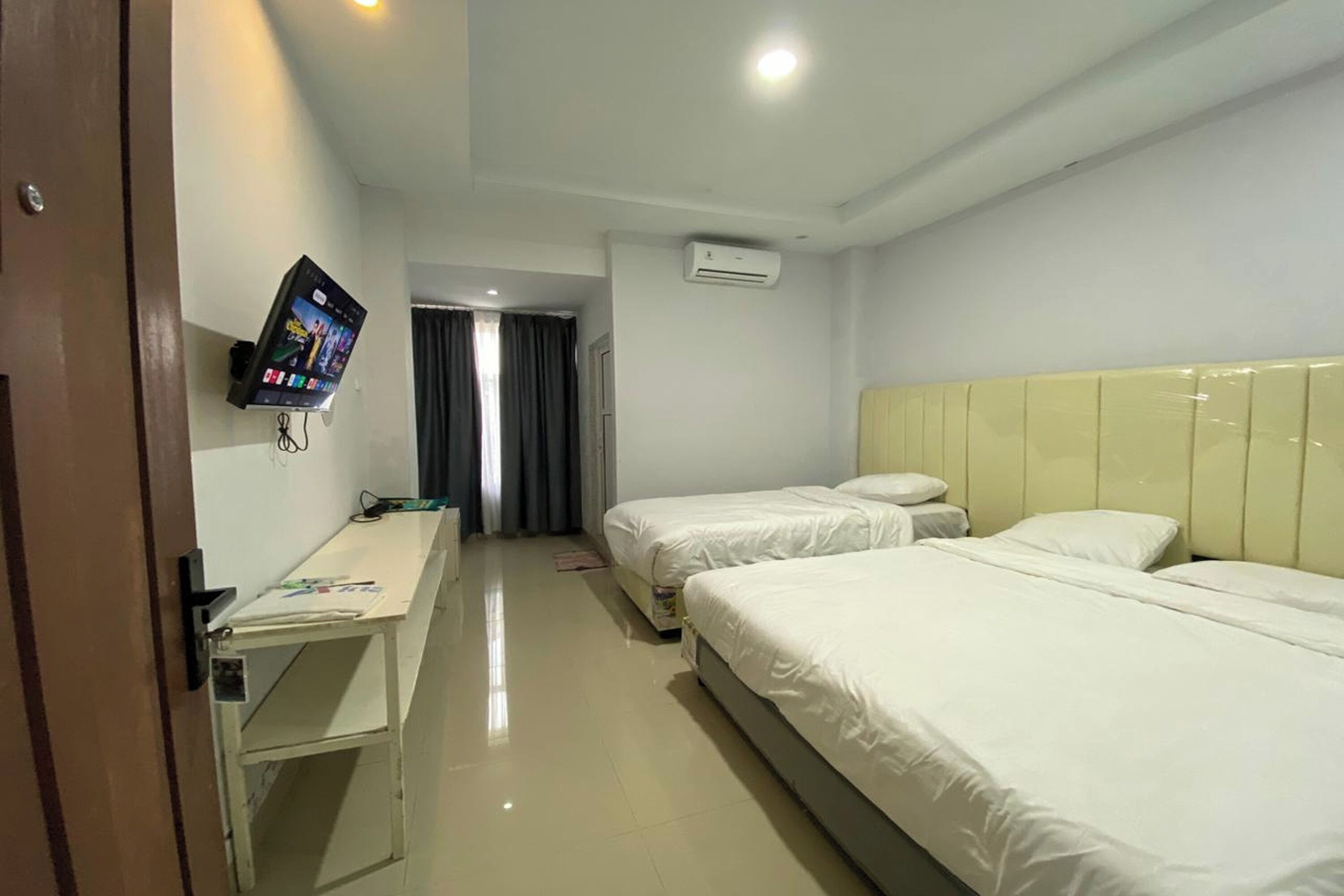 A hotel room with two beds, a desk, a TV mounted on the wall, and an air conditioning unit.