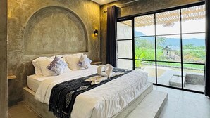 Ein modernes Schlafzimmer mit einem großen Bett, Blick auf Grünflächen und Berge durch ein großes Fenster, und einer Wand mit Betonoptik.