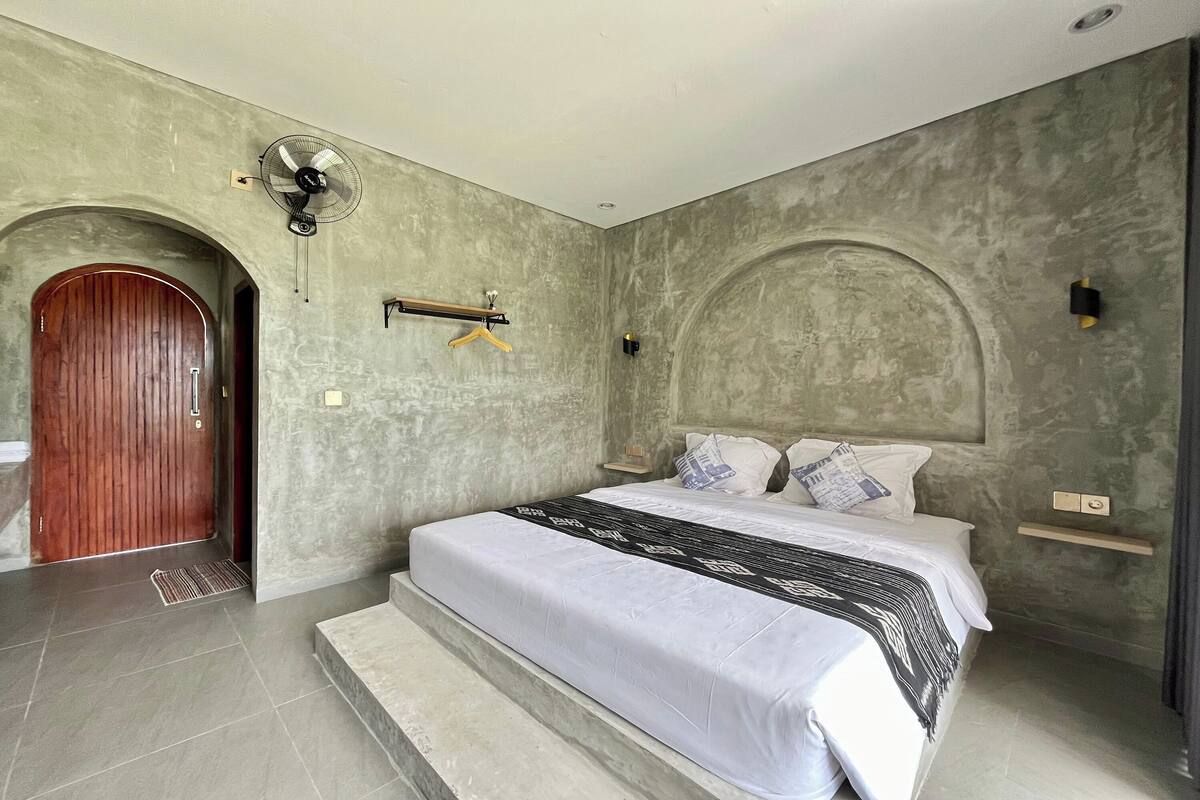 Un dormitorio con cama, una puerta de madera, un ventilador de pared y un perchero.
