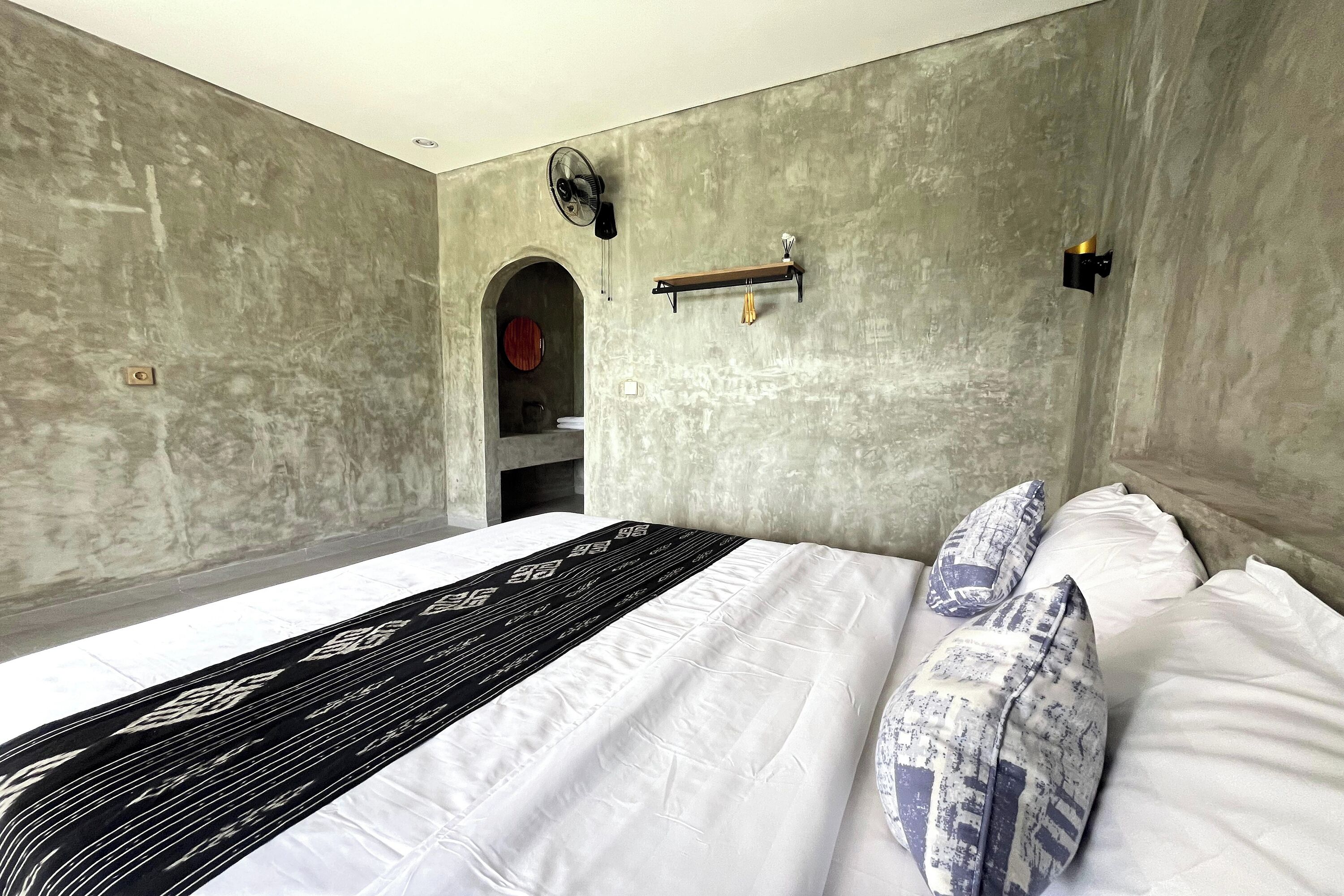 Een slaapkamer met een bed, kussens, een nachtkastje, een ventilator aan de muur en een plank met een lamp.