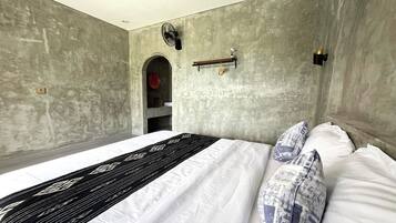 Een slaapkamer met een bed, kussens, een nachtkastje, een ventilator aan de muur en een plank met een lamp.