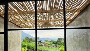 Ein Zimmer mit Betonboden, einer Steinbank und Blick auf eine ländliche Landschaft durch ein Fenster mit Bambusdecke.