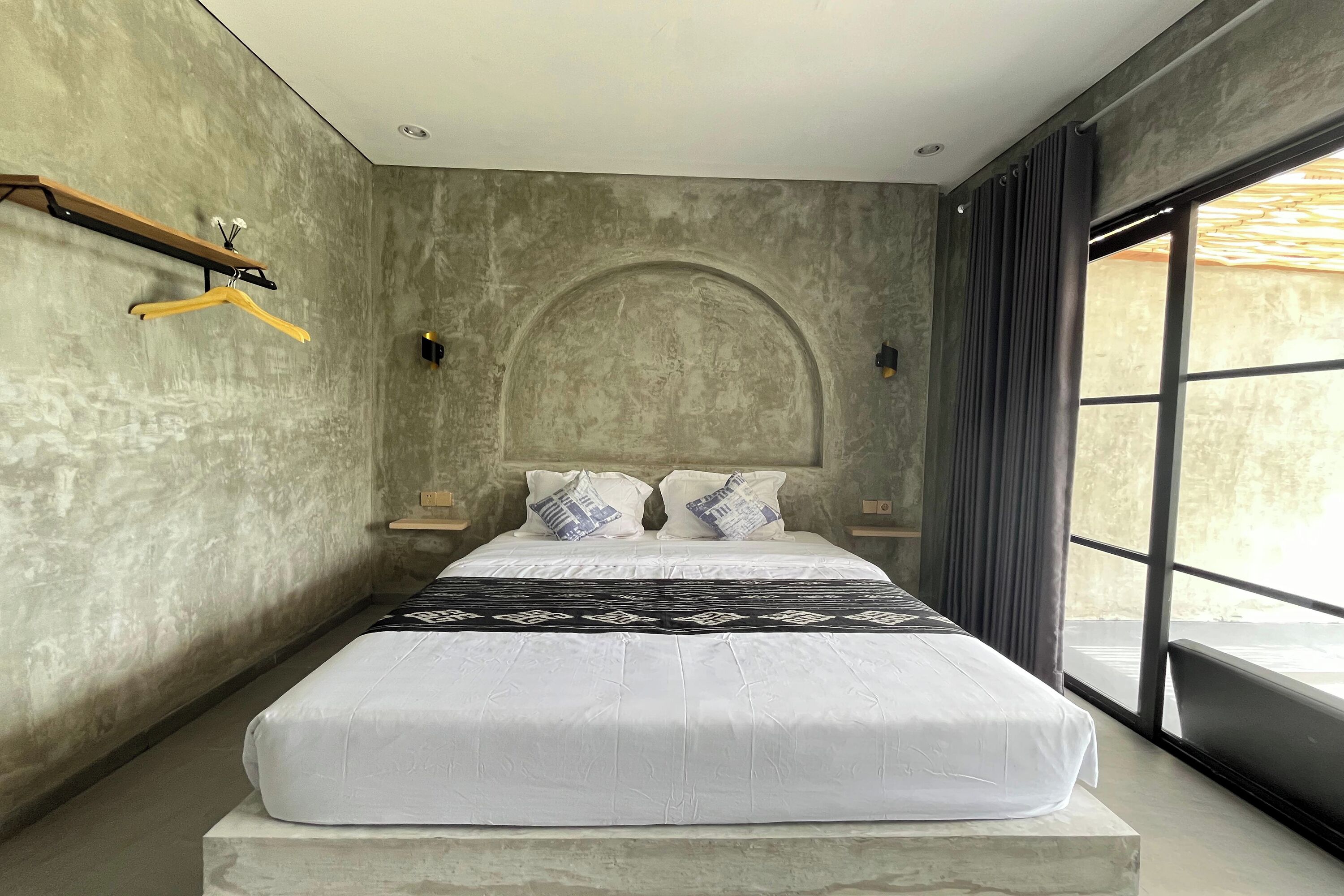 A modern bedroom with a large bed, a wooden shelf with hangers, a headboard, and a large window with curtains.