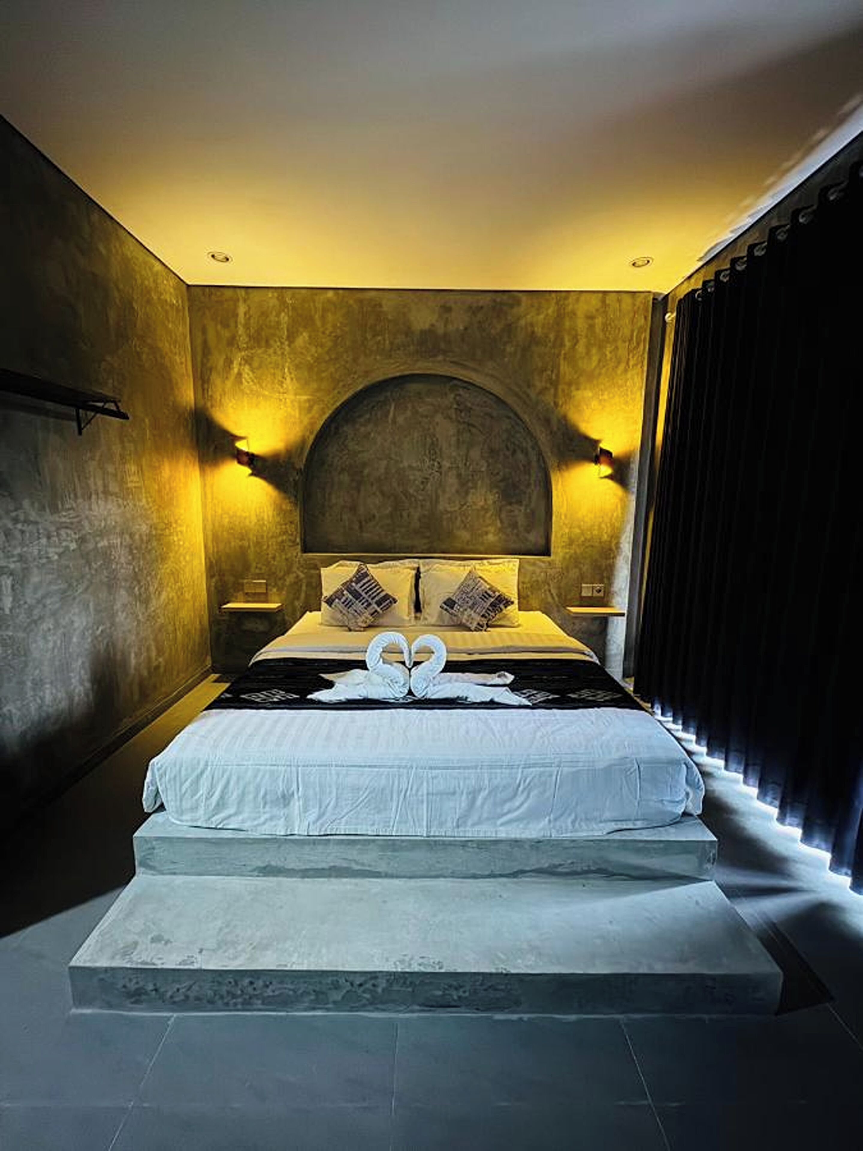 Une chambre à coucher moderne avec un grand lit, deux lampes fixées au mur et un rideau foncé sur le côté droit.