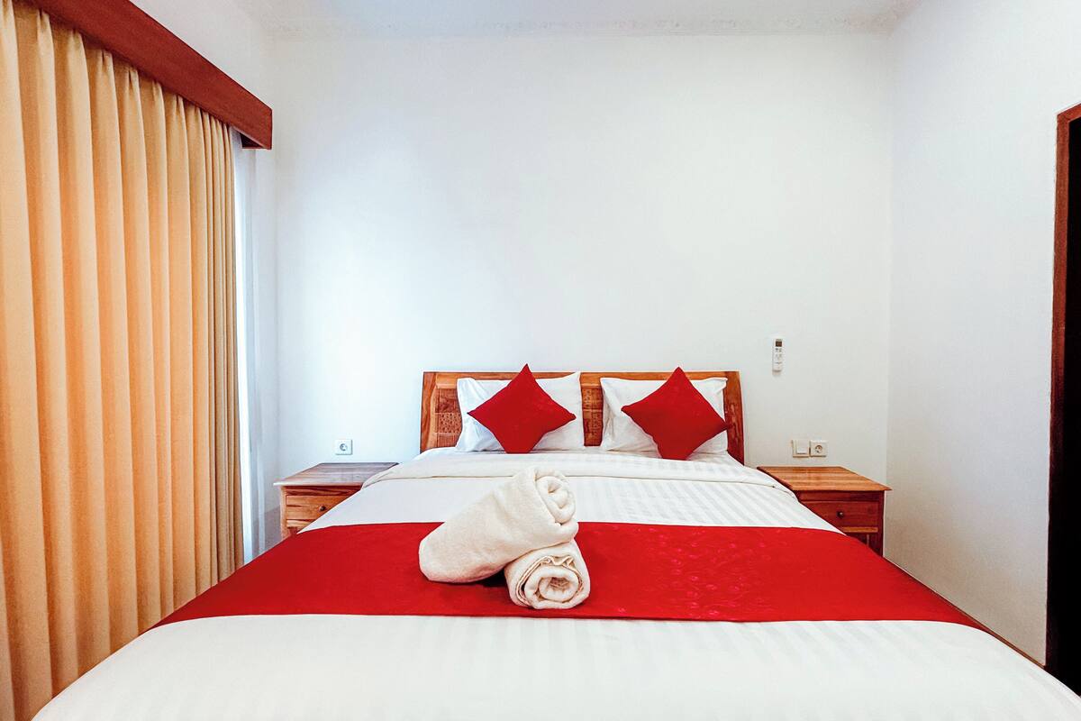 Uma cama bem arrumada com edredom vermelho e branco, duas almofadas vermelhas e uma toalha enrolada sobre a cama.