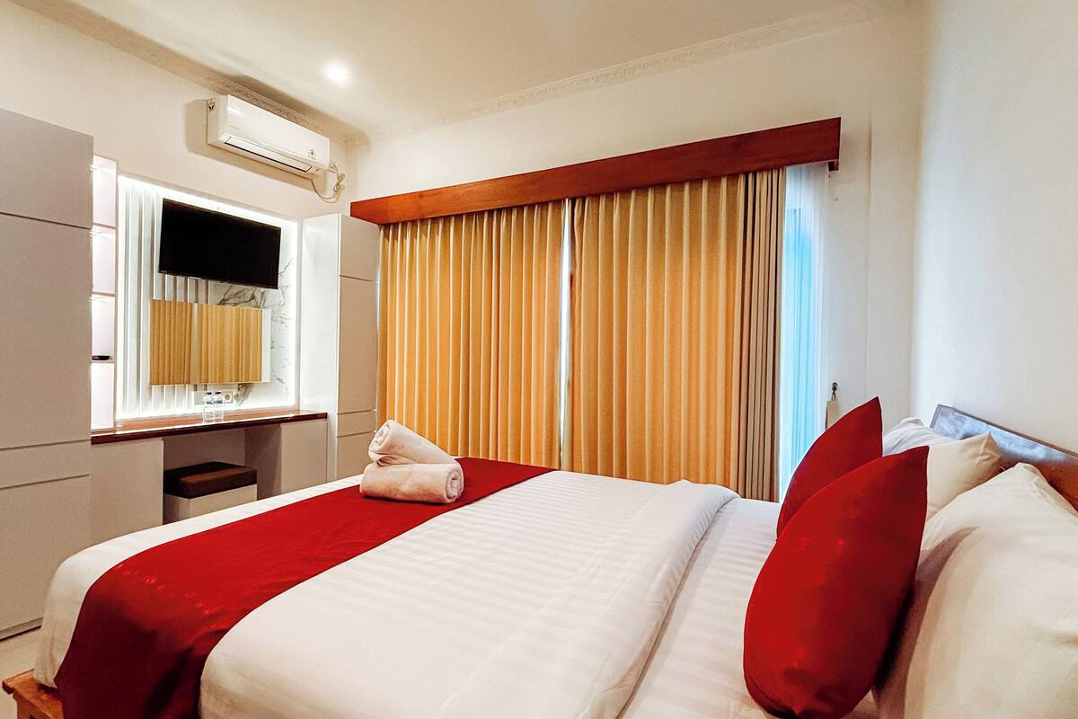 Quarto de hotel com uma cama grande, uma escrivaninha com cadeira, um espelho e uma janela com cortinas.