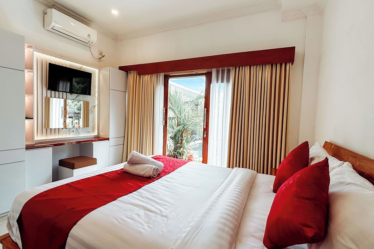 Een hotelkamer met een bed, een aan de muur bevestigde televisie, een raam met gordijnen en uitzicht op palmbomen.