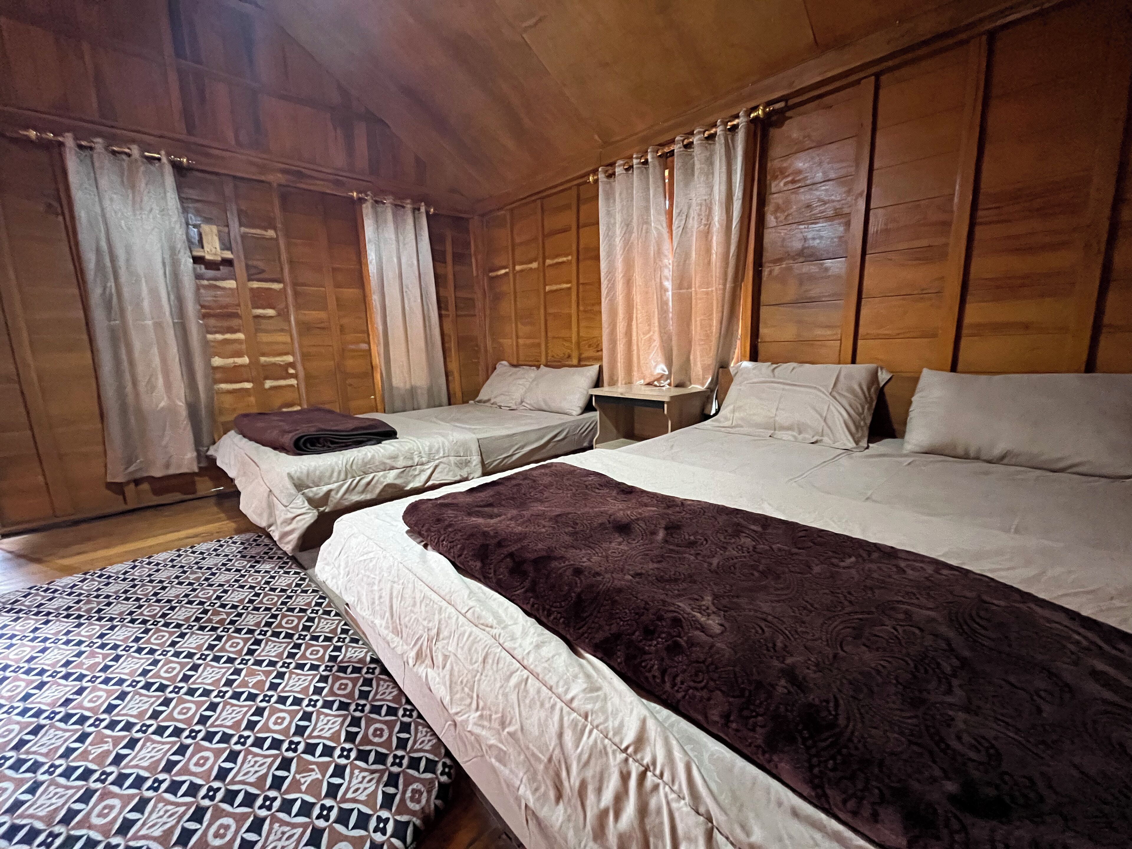 A wooden cabin-style room with two beds, a patterned rug, and curtains.