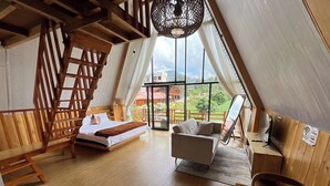 A cozy, wooden-floored room with a bed, a sofa, and a desk. A large window offers a view of greenery outside. - Fifa Cabin Dieng By Fifa Grup (Dieng Kulon)