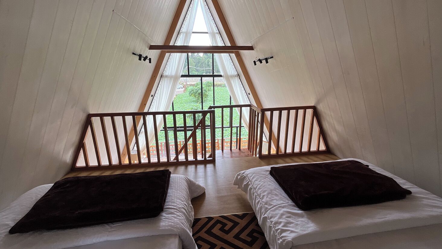 A room with two beds, a wooden railing, and a view of greenery outside.