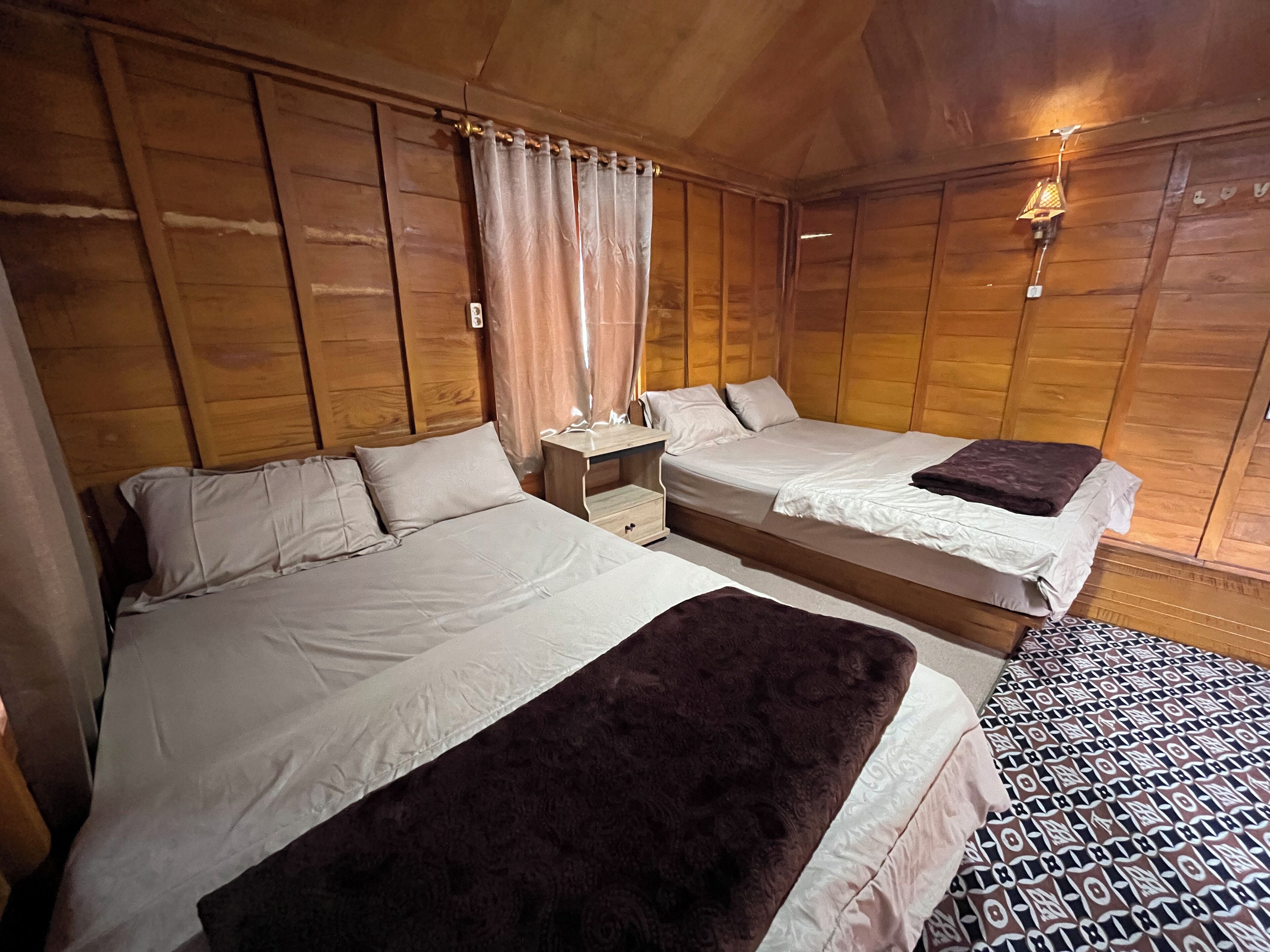 A room with two beds, wooden paneling, a nightstand, and a wall-mounted lamp.