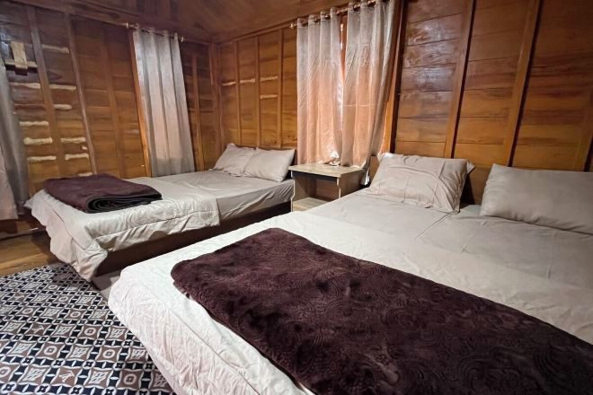 A wooden cabin room with two beds, a small table, and a patterned rug.