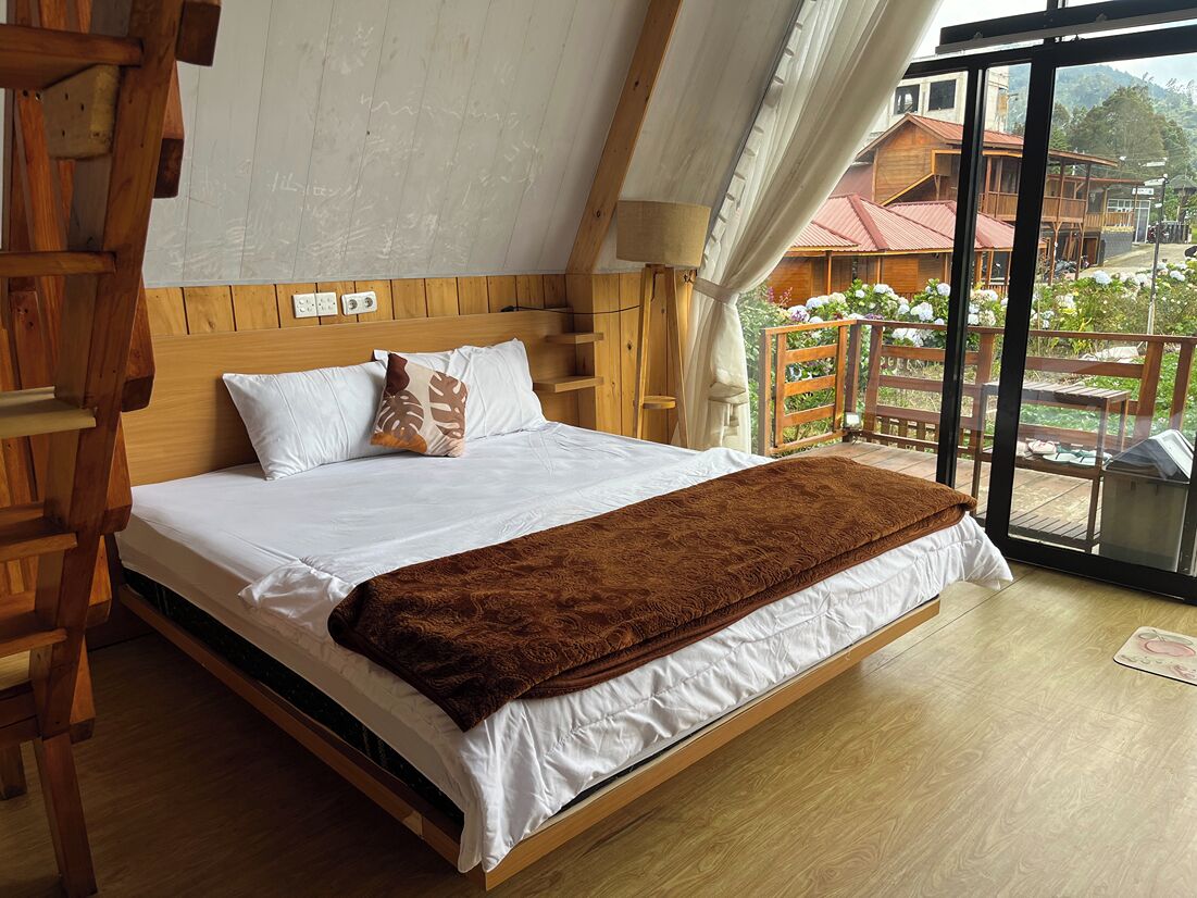 Uma cama de madeira com roupa de cama branca e um cobertor castanho, uma escada de madeira e uma janela grande com vista para um edifício e vegetação lá fora.