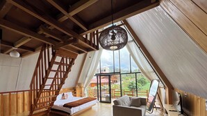 A loft-style bedroom with a wooden staircase, a bed, a sofa, and a large window offering a view of the outdoors. - Fifa Cabin Dieng By Fifa Grup (Dieng Kulon)