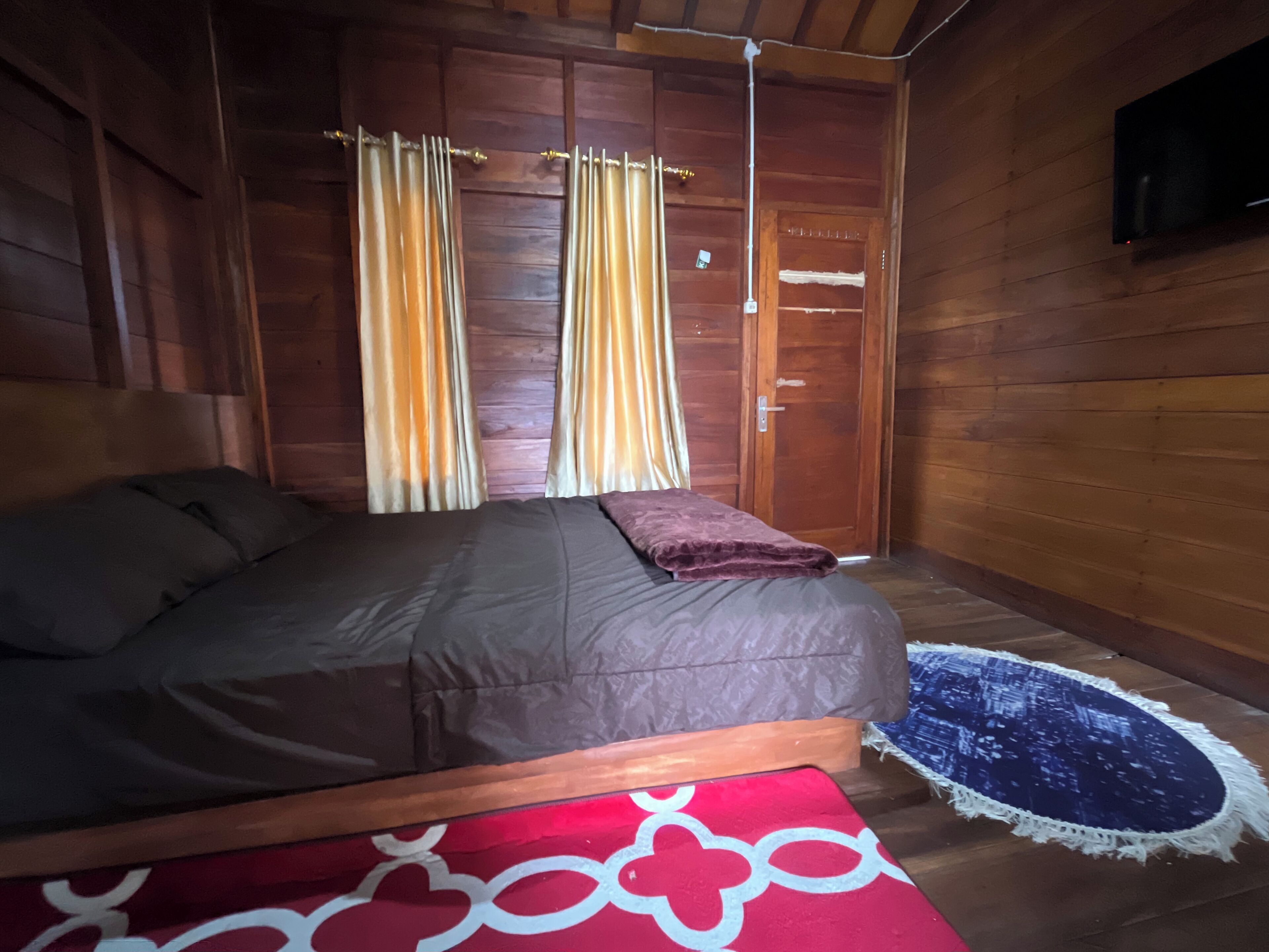 Une chambre dans un chalet en bois, avec un lit, des rideaux, une porte et un téléviseur fixé au mur.