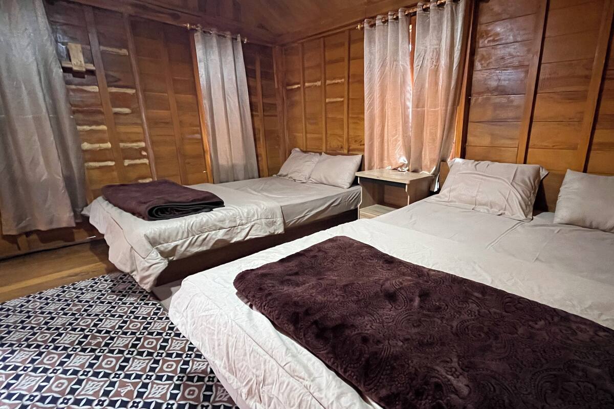A wooden cabin-style room with two beds, a small table, and a patterned rug.
