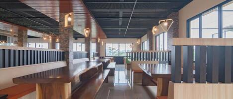A spacious restaurant with high wooden benches and tables, a modern ceiling design, and large windows allowing natural light.
