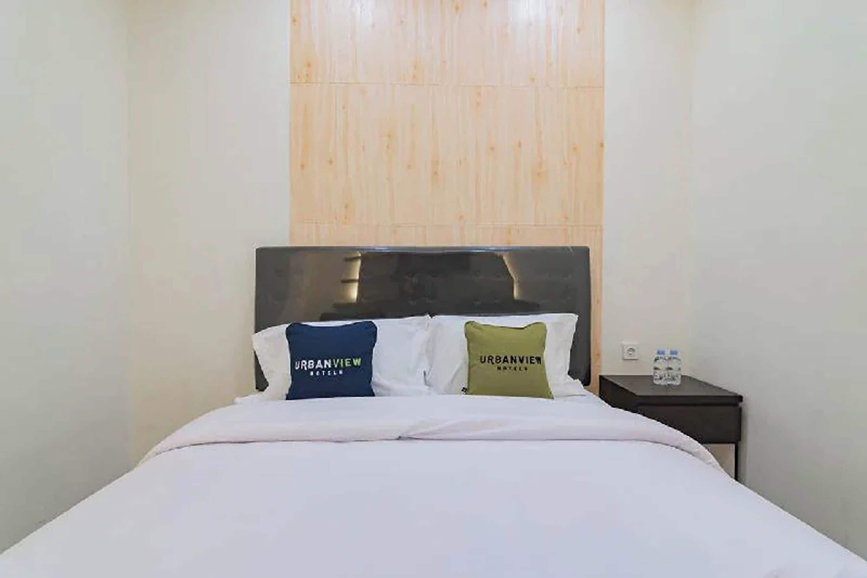 A hotel room with a bed, two pillows with 'URBANVIEW HOTEL' text, a nightstand with a water bottle, and a wooden panel wall.