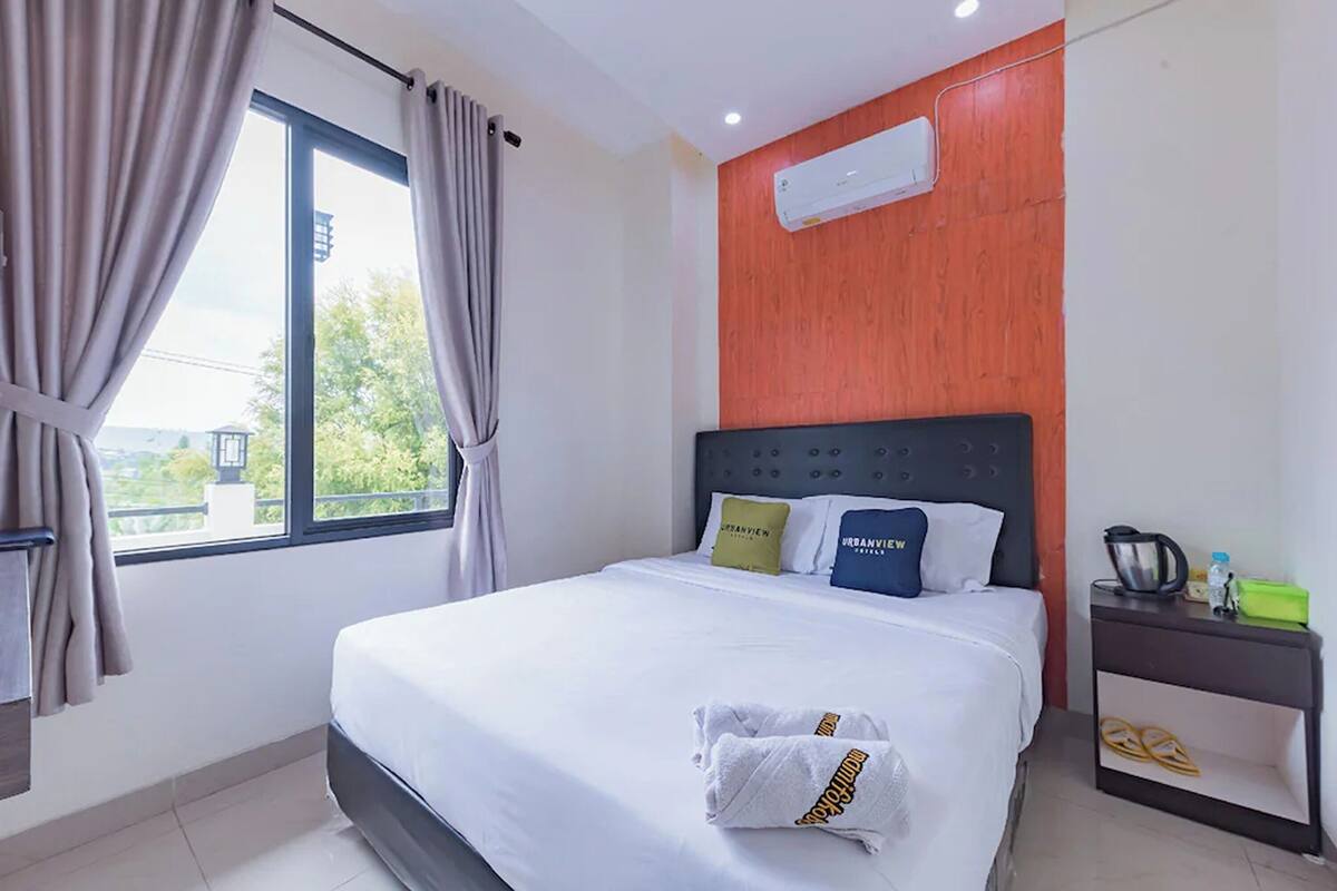 Habitació d'hotel amb un llit, una petita taula de nit, aire condicionat i una finestra amb cortines.